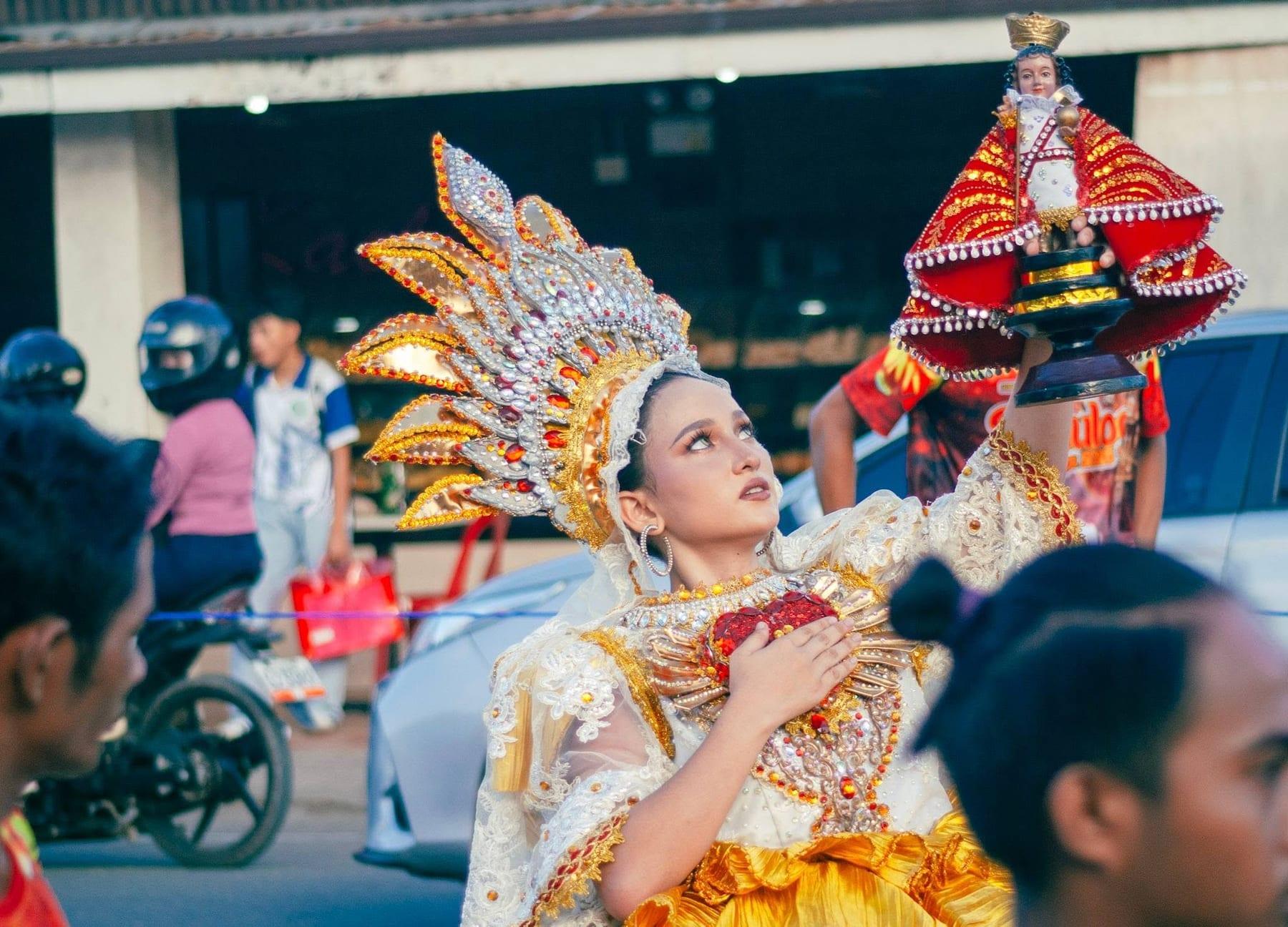 Woman in an ornate festival costume holding a Santo Niño statue aloft during a street celebration