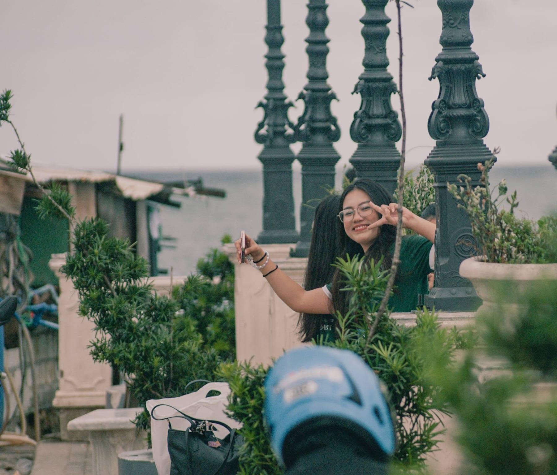 Candid lifestyle photograph of a smiling young woman making a peace sign along a coastal promenade