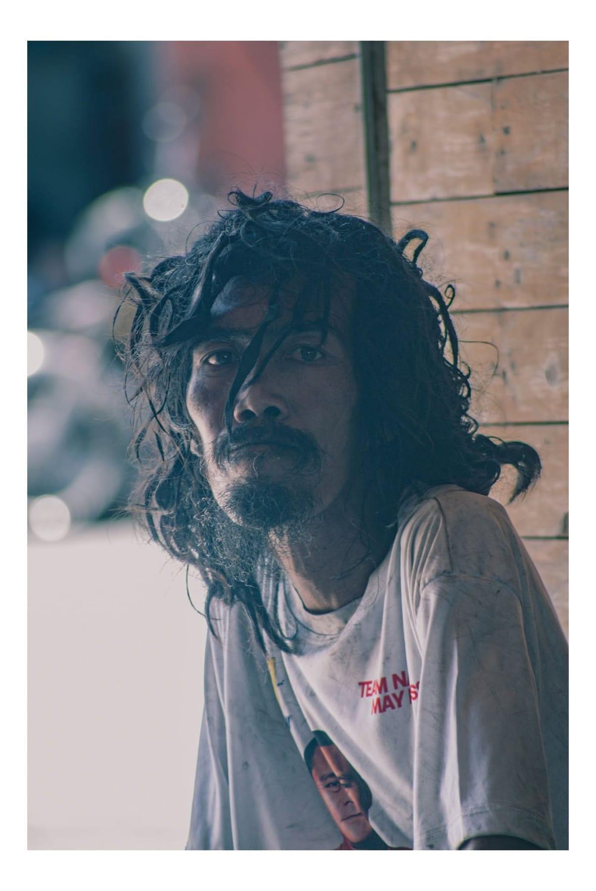 Documentary-style portrait of a man with long dark hair leaning against a wooden wall